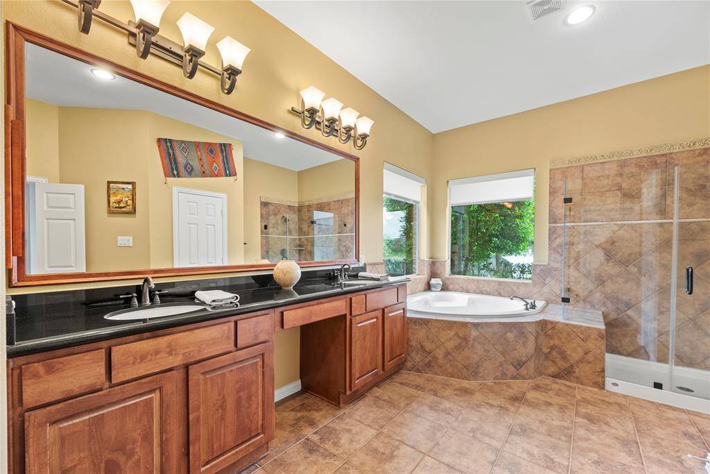 This spacious primary bathroom features a double vanity and sinks with granite countertop, and a large mirror. It includes a luxurious corner bathtub and a separate glass-enclosed shower which includes a bench seat.