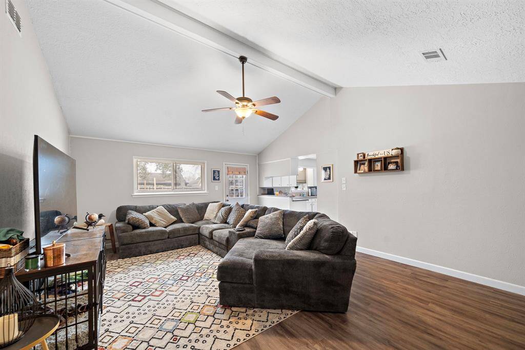 This inviting living room features high ceilings with exposed beams and a ceiling fan for comfort. The space is bright with natural light. The room has a warm, neutral color palette and wood-looking flooring, creating a cozy atmosphere.