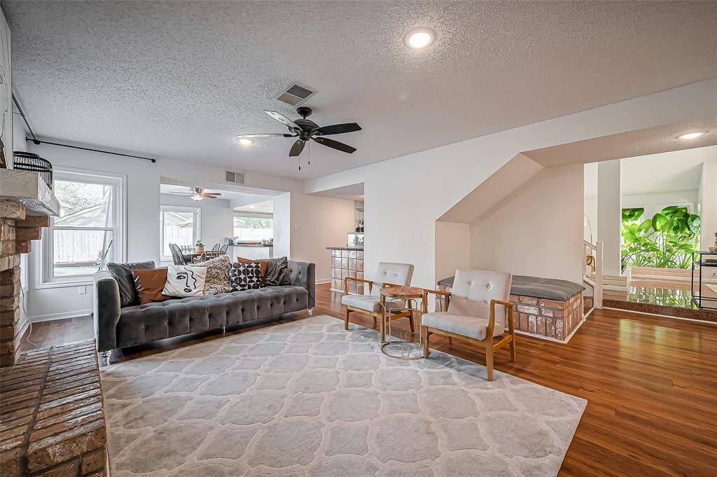 This inviting living room features a cozy seating area with a plush sofa and modern chairs, a ceiling fan, and warm wood flooring. Natural light streams through large windows, highlighting the spacious layout. A neutral-toned rug and a brick accent wall add charm and character.