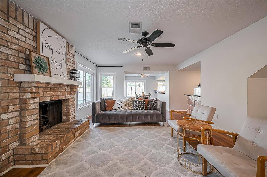 This inviting living room features a cozy brick fireplace, plush seating, and large windows that let in plenty of natural light. The open layout leads into a dining area, creating a seamless flow for entertaining. Ceiling fans and modern decor add to the comfortable, stylish ambiance.