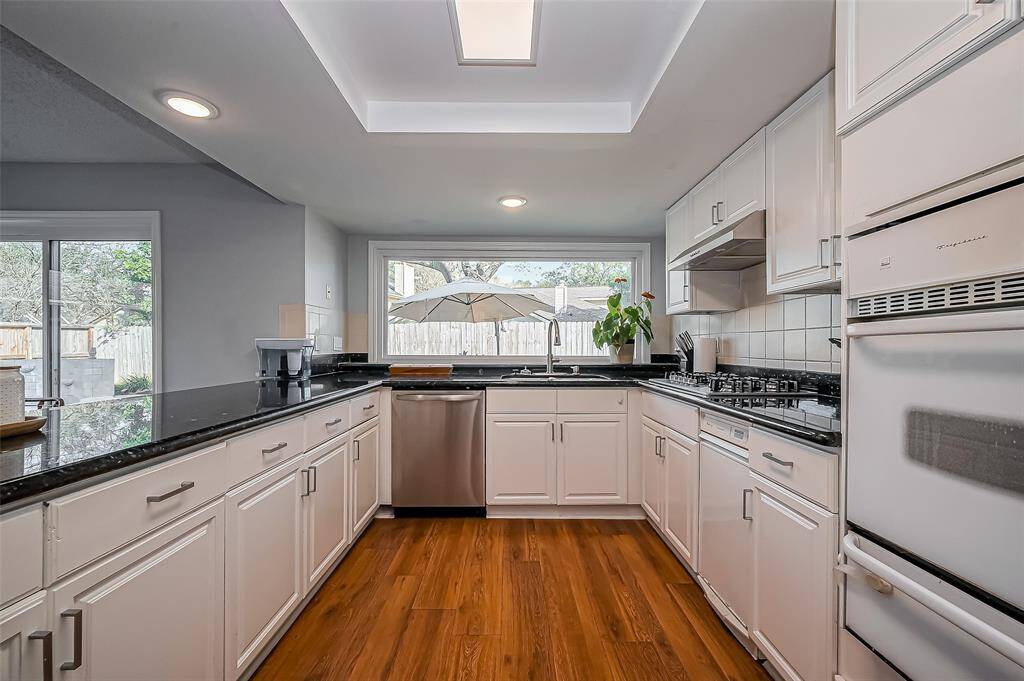 This modern kitchen features sleek white cabinetry, stainless steel appliances, and a spacious layout. It has a large window offering plenty of natural light and a view of the backyard, along with a sliding door for easy outdoor access. The dark countertops provide a stylish contrast to the wood flooring.