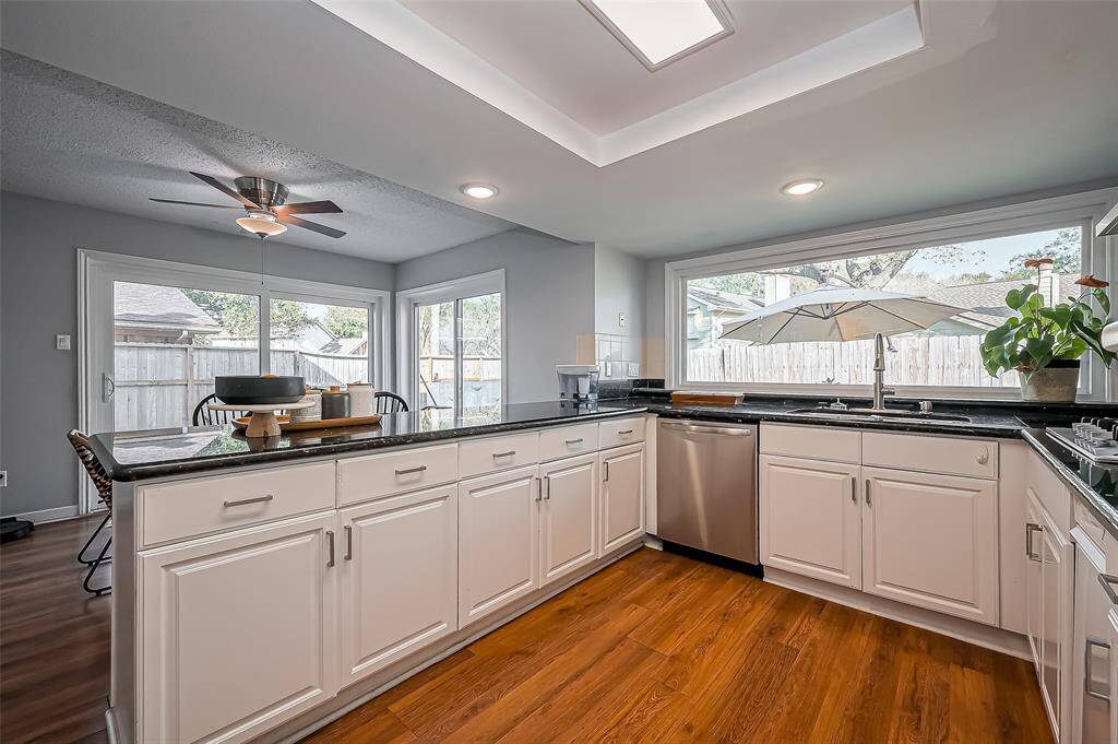 This kitchen features modern white cabinetry, dark countertops, and stainless steel appliances. It has a bright, open layout with large windows and sliding glass doors leading to the backyard. The space includes a dining area with a ceiling fan and wood flooring, enhancing the warm and inviting atmosphere.