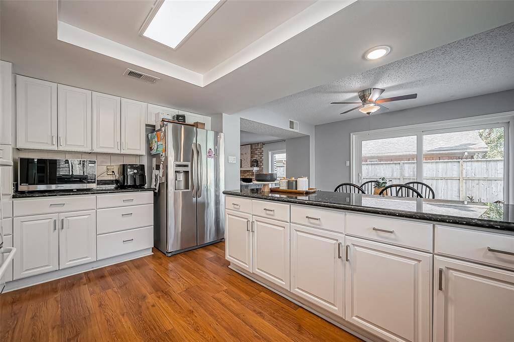 This kitchen features modern stainless steel appliances, spacious white cabinetry, and sleek black countertops. It opens to a dining area with a ceiling fan and large sliding glass doors, providing ample natural light and access to the backyard. The hardwood floors add warmth and elegance to the space.