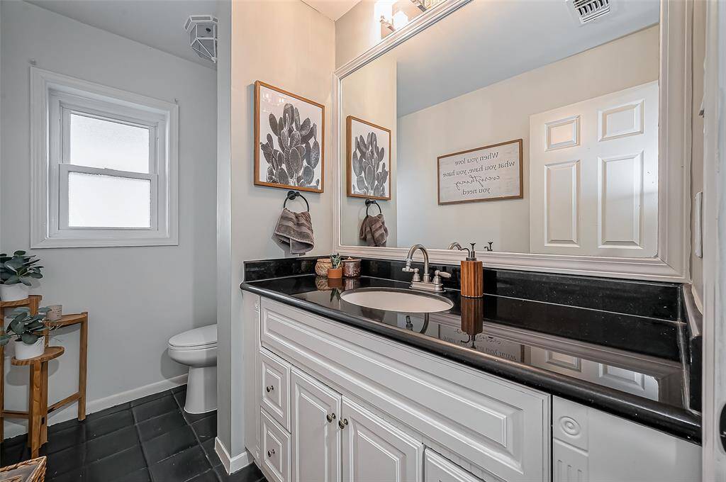 This bathroom features a sleek design with a black countertop and white cabinetry. It includes a large mirror, modern fixtures, tasteful wall art, and natural light from a frosted window. The space is complemented by decorative plants and neutral tones.