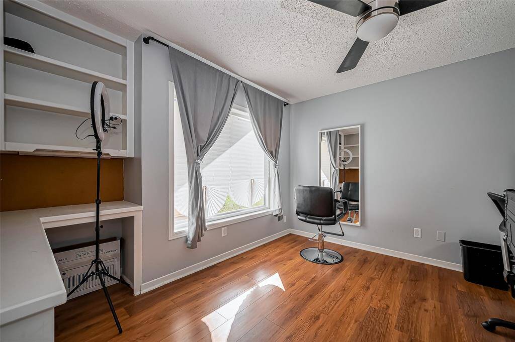 This modern, well-lit room features hardwood floors, a large window with gray curtains, a ceiling fan, and built-in shelving. Perfect for a home office or creative space.
