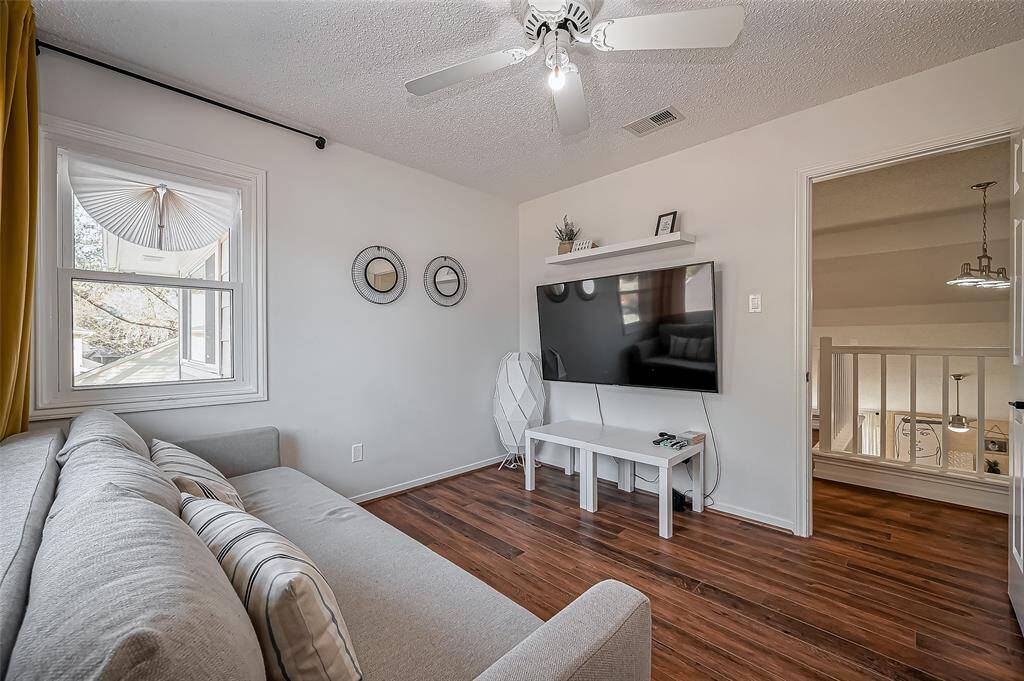 This cozy room features a comfortable sofa, a wall-mounted TV, and a ceiling fan. The space is well-lit with natural light from a window, and has stylish hardwood flooring. A small shelf adds a decorative touch above the TV.