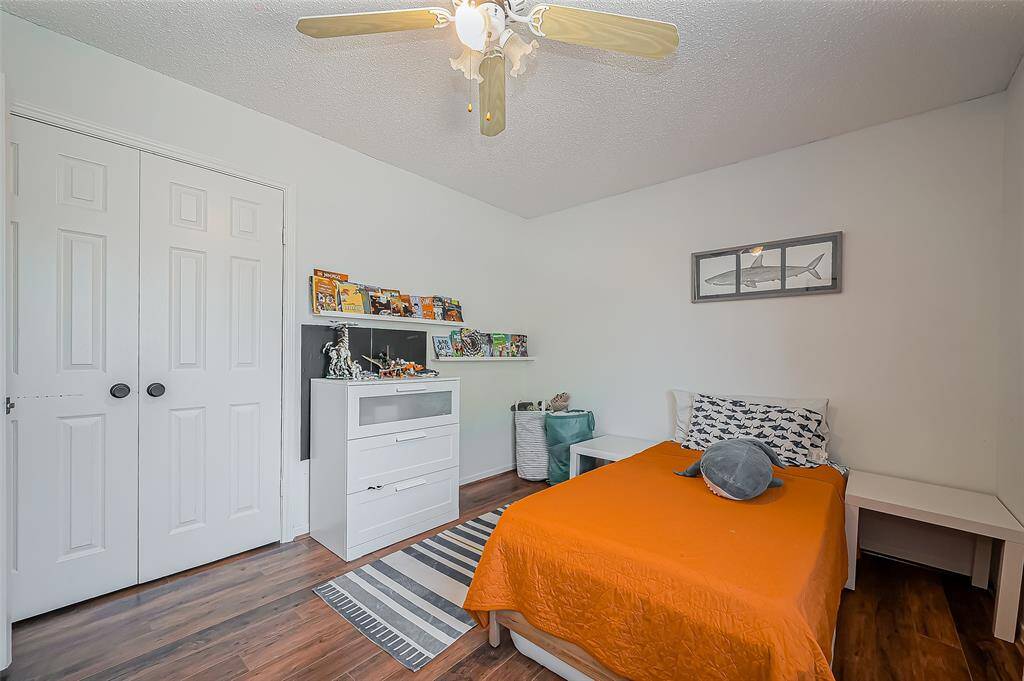 This cozy bedroom features a warm wooden floor, a ceiling fan, and a bright orange bedspread. It includes a spacious closet with double doors, a white dresser with shelving above, and a simple, modern decor that offers a clean and inviting atmosphere.