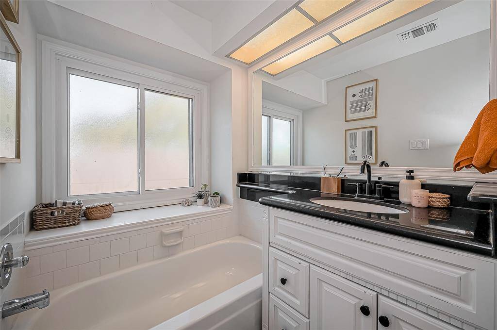 This bathroom features a clean, modern design with a white tub, black countertop, and ample lighting. A large window provides natural light, and there are decorative elements like framed art and small plants for a touch of warmth.