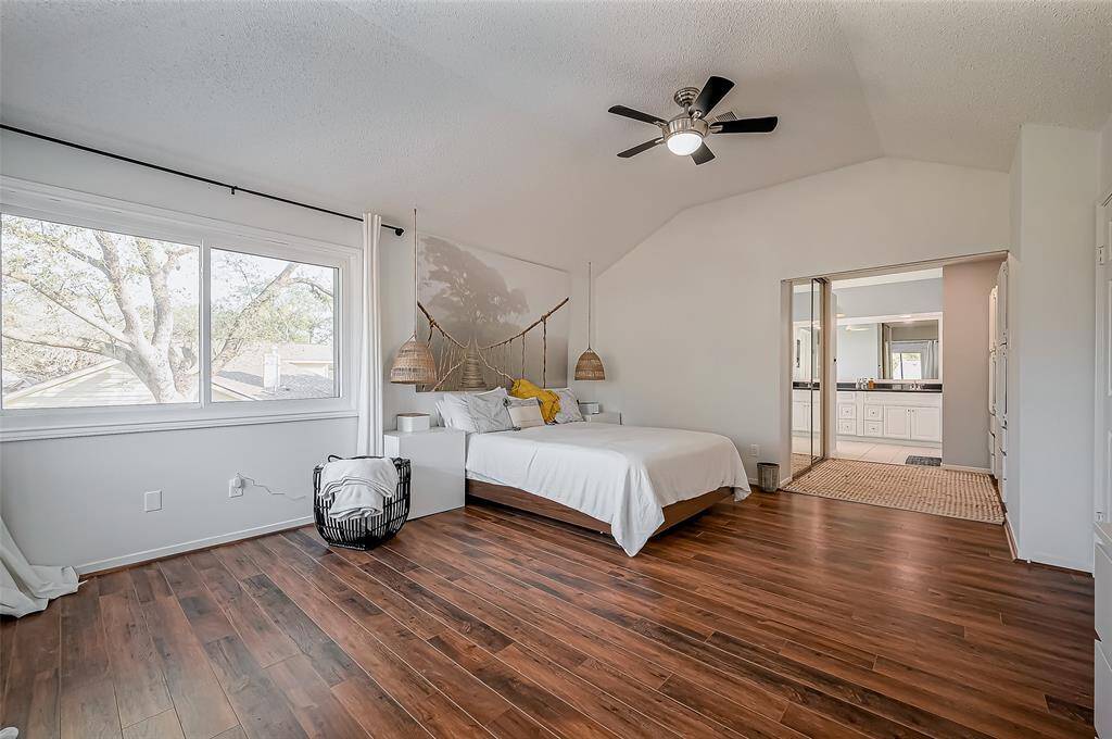 This spacious bedroom features warm wood flooring, a large window with ample natural light, and a vaulted ceiling with a modern ceiling fan. It opens into a bright en-suite bathroom, providing a seamless flow. Perfect for comfort and style.