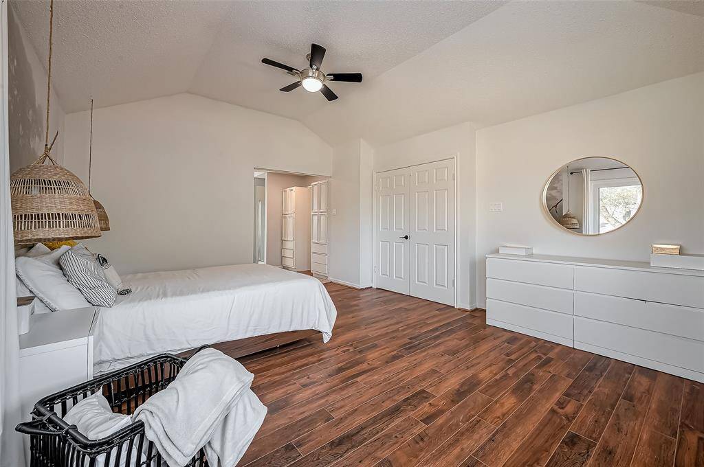 Bright and spacious bedroom with vaulted ceilings, featuring hardwood floors and a modern ceiling fan. It includes a cozy bed, a wicker pendant light, a white dresser with a round mirror, and a large closet. Natural light streams through a window, creating an inviting atmosphere.