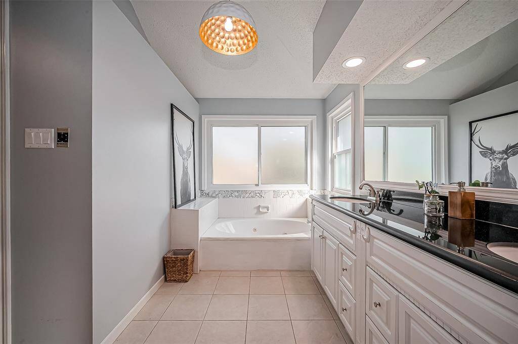Bright and inviting bathroom featuring a large soaking tub, dual-sink vanity with black countertop, and ample natural light from frosted windows. Neutral tones and modern lighting add to the elegant ambiance.