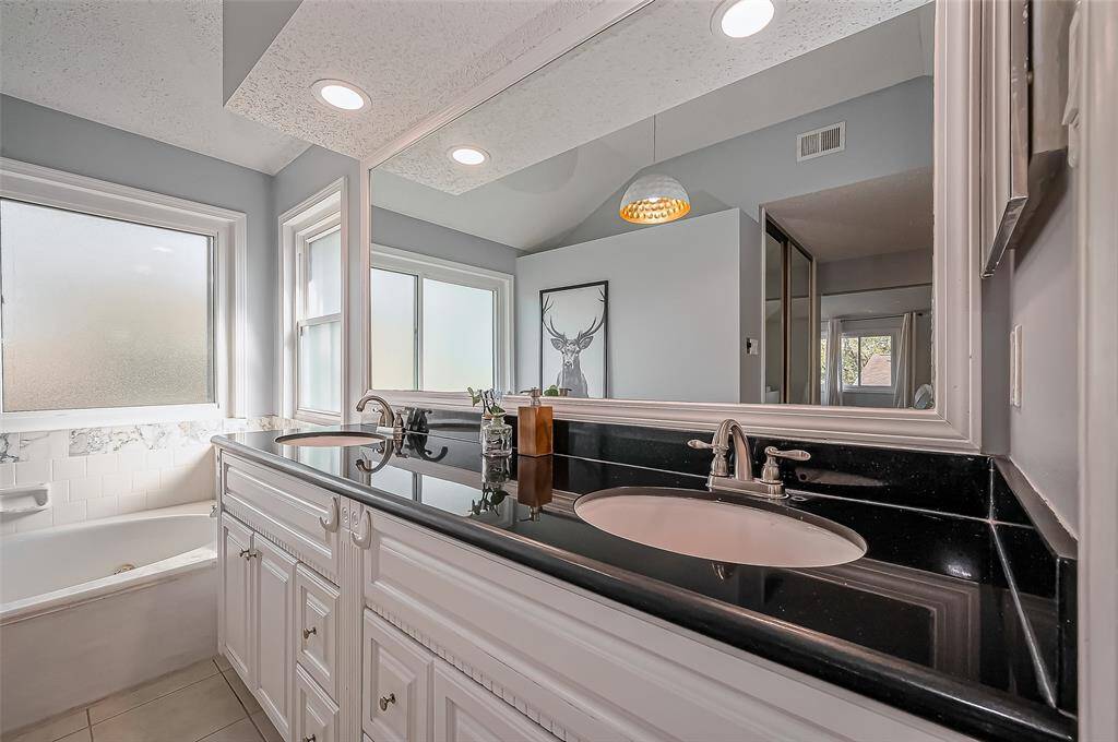 This bathroom features a modern double vanity with sleek black countertops and white cabinetry. It includes a bathtub with frosted windows for privacy, recessed lighting, and stylish decor, creating a bright and contemporary space.