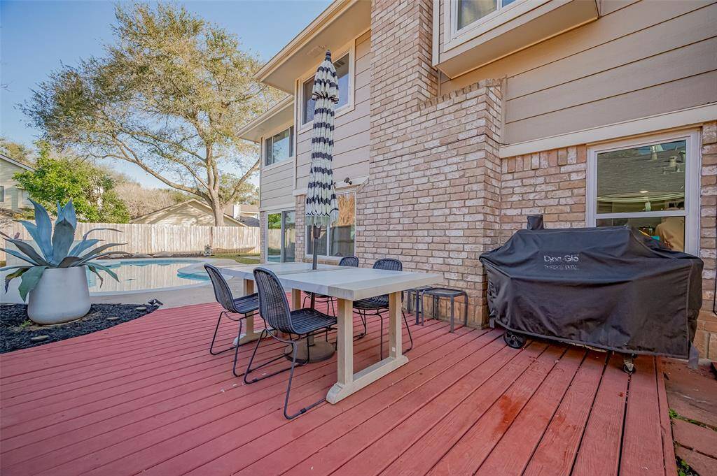 This outdoor space features a cozy red deck with a modern dining set and a covered grill. It's adjacent to a pool, surrounded by lush greenery and a privacy fence, perfect for entertaining or relaxing.