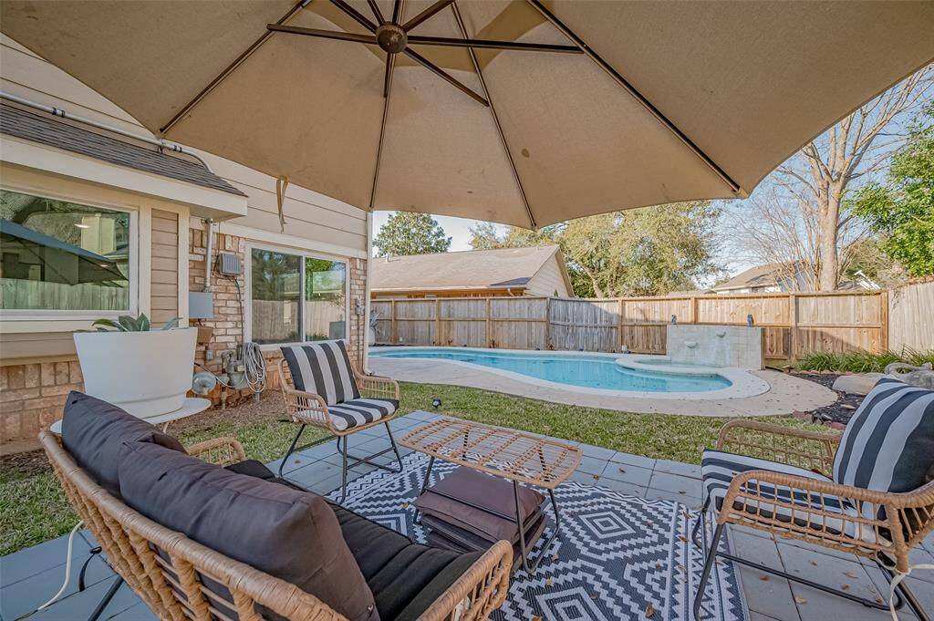 This inviting backyard features a cozy patio with comfortable seating and a large umbrella, overlooking a refreshing in-ground pool. The area is surrounded by a wooden privacy fence and well-maintained landscaping, providing a perfect spot for relaxation and outdoor entertaining.