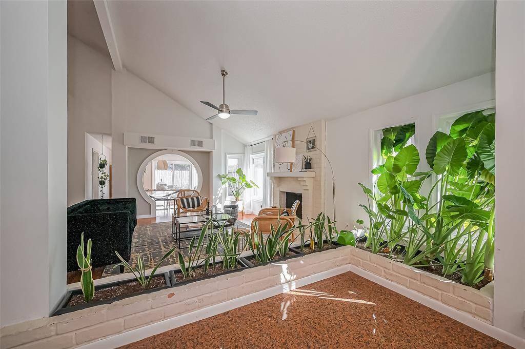 This spacious living area features a vaulted ceiling, modern fan, and a cozy fireplace. Large windows and lush indoor plants create a bright and inviting atmosphere, while the open layout seamlessly connects to the dining area.