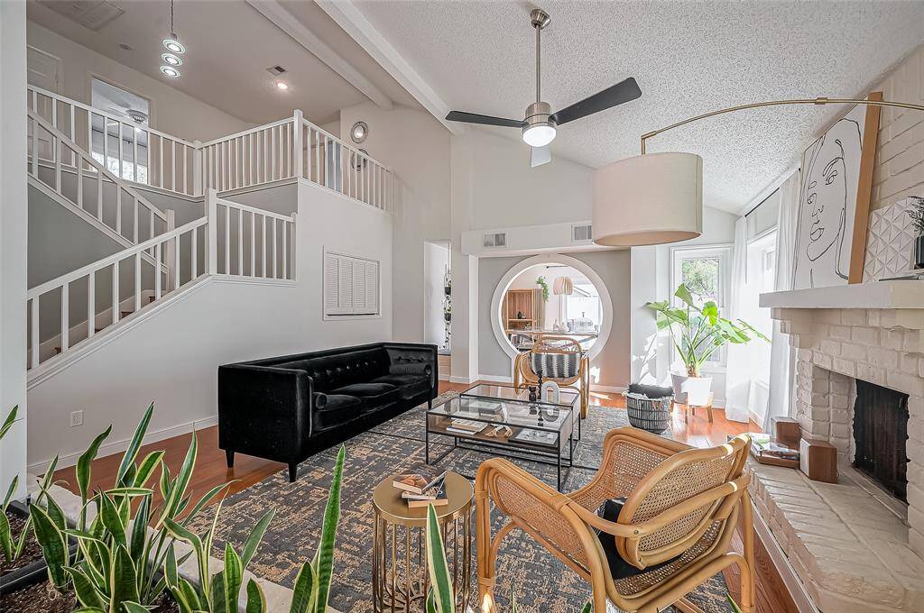 This modern living room features an open layout with high ceilings, a sleek black sofa, and a cozy fireplace. It has stylish decor, including a large circular mirror and contemporary artwork. Natural light floods in through large windows, highlighting the elegant staircase and warm wood flooring.