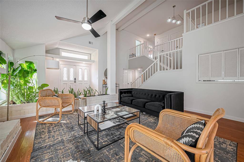 This spacious living room features high ceilings, a modern ceiling fan, and abundant natural light. The room is elegantly furnished with a sleek black sofa and stylish wicker chairs. A glass coffee table complements the contemporary décor, while lush indoor plants add a touch of greenery. The open staircase and white walls enhance the airy feel.