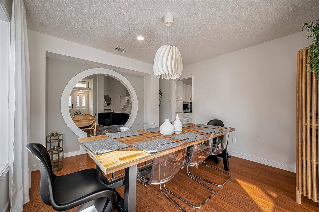 This photo showcases a modern dining room with a sleek wooden table and clear chairs. It features a stylish pendant light and a large circular mirror on the wall. The space is bright with natural light from a nearby window, and the hardwood floor adds warmth.