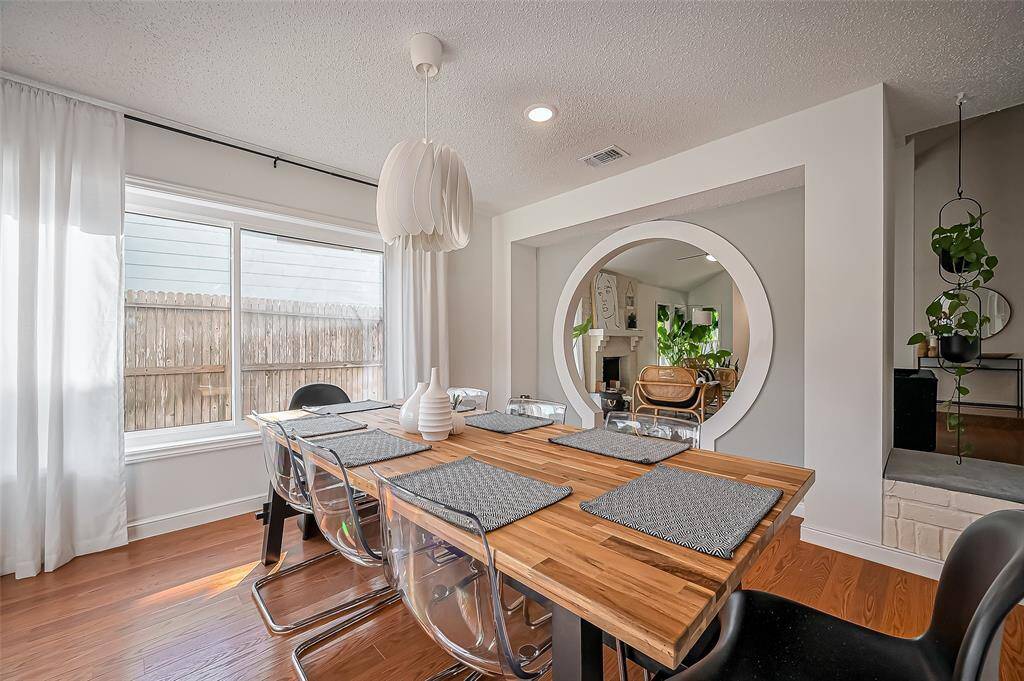 Bright and modern dining area with a wooden table, clear chairs, and a stylish pendant light. Features a large window with sheer curtains and a circular mirror adding depth. The space connects to a cozy living area with plants, offering a welcoming atmosphere.