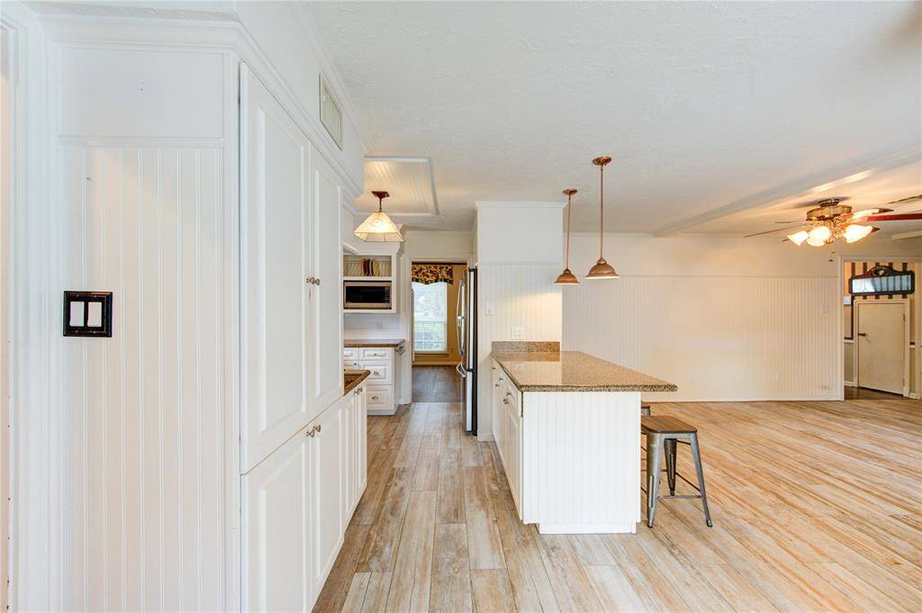 The kitchen features crisp white cabinetry, granite countertops, a spacious island with bar seating, and stylish pendant lighting—all flowing into a bright and open living area with wood-look tile flooring.