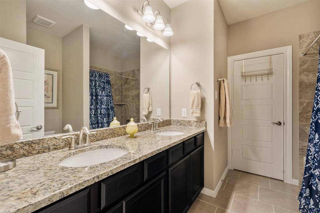 This photo showcases a modern bathroom with dual sinks featuring granite countertops. It includes a tiled shower, and neutral-toned walls and floors. Serves as an ensuite & secondary bath for guests.