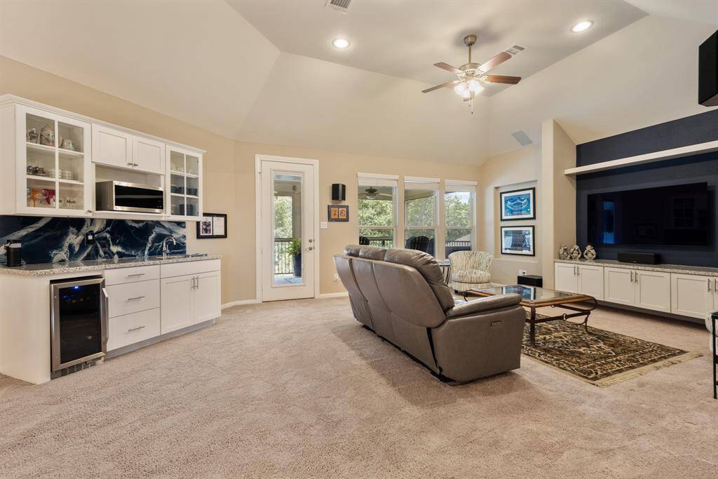 This large game room features a vaulted ceiling, plush carpeting, and abundant natural light from large windows. It includes a spacious area facing a built-in entertainment center. Added TV entertainment wall with granite countertop, floating shelving and cabinets with undermount lighting, and art niche. Wet bar with granite countertop, custom backsplash, wine fridge, microwave, lower storage and upper glass cabinets. *TV excluded.