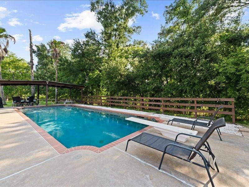 The serene backyard with the pool surrounded by lush greenery, features a diving board and lounge chairs on a spacious patio.