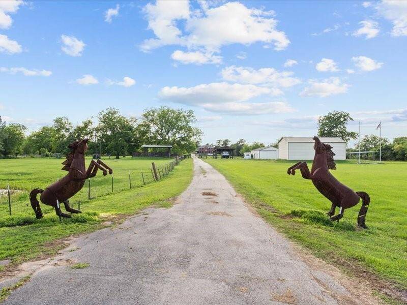 Charming rural property with a long driveway flanked by two horse sculptures, leading to a spacious, open area with green fields and a large outbuilding, perfect for country living.