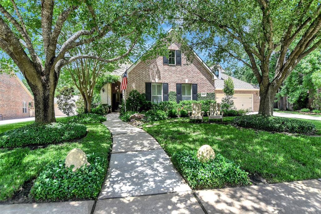 Beautiful brick home with a welcoming walkway, surrounded by lush greenery and mature trees. Features a well-maintained lawn, cozy porch seating, and a spacious driveway leading to a three-car garage.