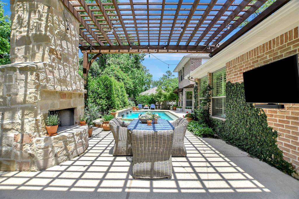 This inviting outdoor space features a pergola-covered patio with a stone wood burning fireplace,  and wall-mounted TV. It overlooks a sparkling pool, surrounded by lush greenery, offering a perfect setting for relaxation and entertainment.