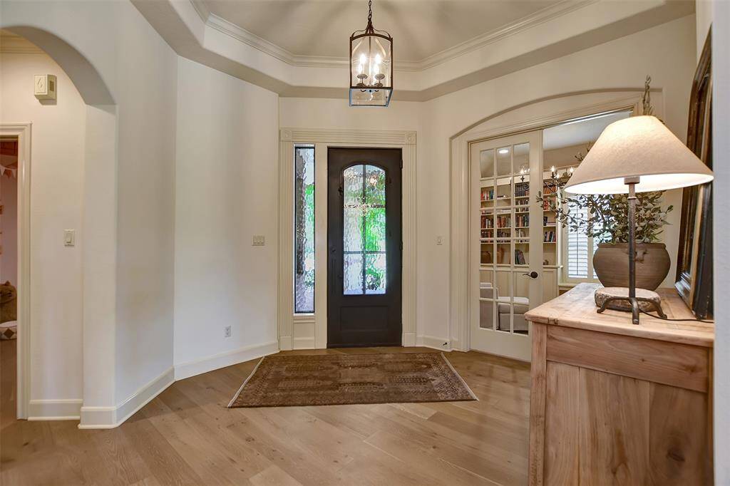 This inviting entryway features a stylish iron front door with glass panels, allowing natural light to flow in. The space includes elegant engineered wood flooring and a cozy ambiance enhanced by a decorative fixture. Adjacent is a study with French doors, offering a glimpse of a well-organized office or library.