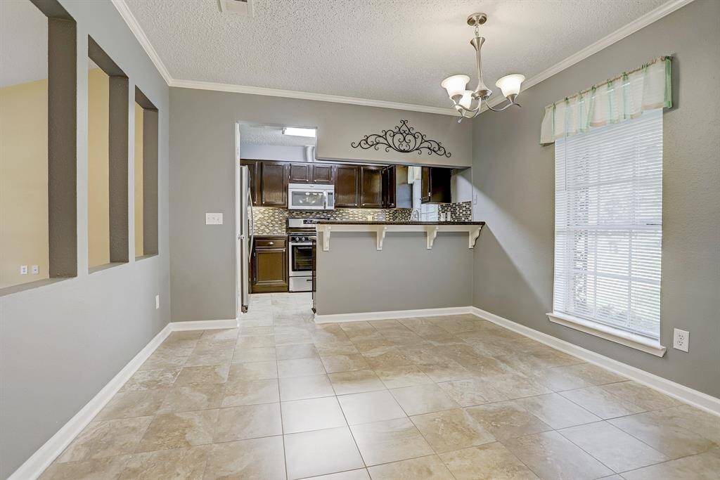 Bright dining area with tile flooring, open wall cutouts, and a pass-through to a modern kitchen featuring dark cabinetry. A large window and elegant light fixture add charm and natural light.