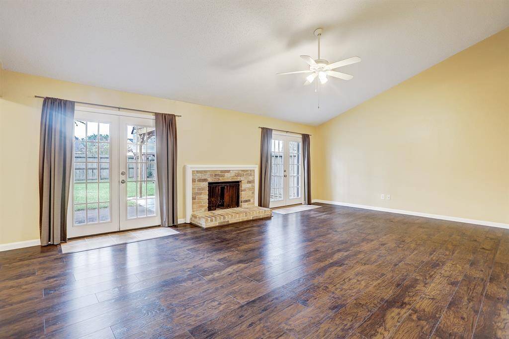 Spacious living room with high ceilings, featuring a cozy brick fireplace and dark hardwood floors. The room is brightened by natural light from two sets of French doors, leading to a fenced backyard.