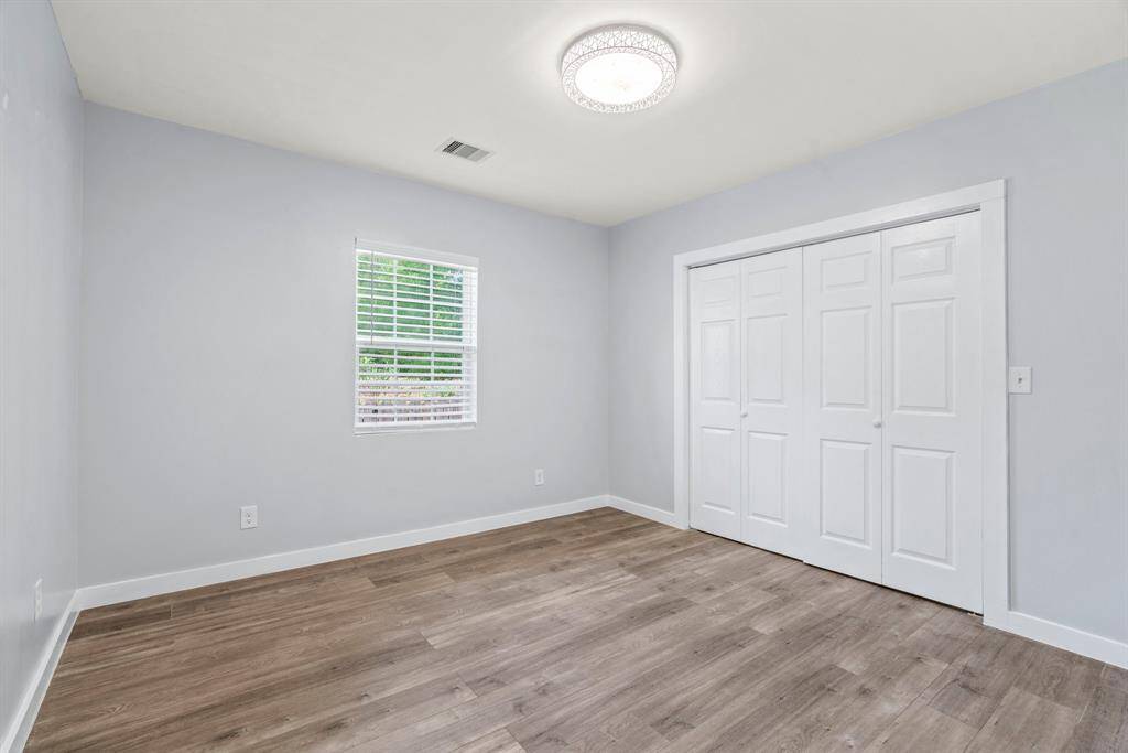 Bright and airy bedroom with light gray walls and wood-style flooring. Features a modern ceiling light, a window with white blinds, and a spacious white double-door closet.