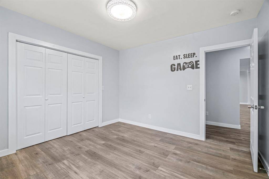 This room features fresh light gray walls, a modern ceiling light fixture, and wood-look flooring. It includes a double-door closet and a playful "Eat. Sleep. Game." wall decal, ideal for a bedroom or gaming room.
