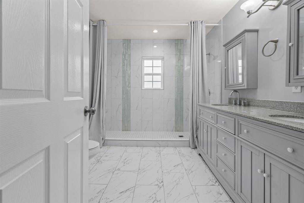 This bathroom features a spacious design with elegant marble-patterned tiles, a large vanity with dual sinks, and a walk-in shower. Soft gray cabinetry and a window provide a modern and airy feel.