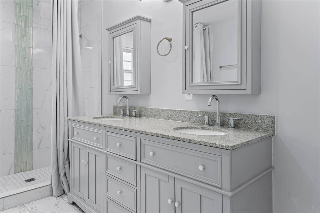This photo showcases a modern bathroom with a double vanity featuring granite countertops and elegant light gray cabinetry. It includes two mirrors and a stylish, tiled shower area with an accent strip. The design is clean and sophisticated, ideal for a contemporary home.