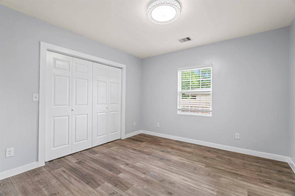 This photo shows a bright, minimalist bedroom with light gray walls, hardwood flooring, a modern ceiling light, a window with blinds, and a double-door closet.