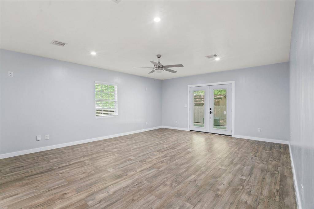 This spacious room features sleek wood flooring, soft gray walls, and ample natural light from a window and glass double doors. A ceiling fan enhances comfort, making it ideal for a modern living area.