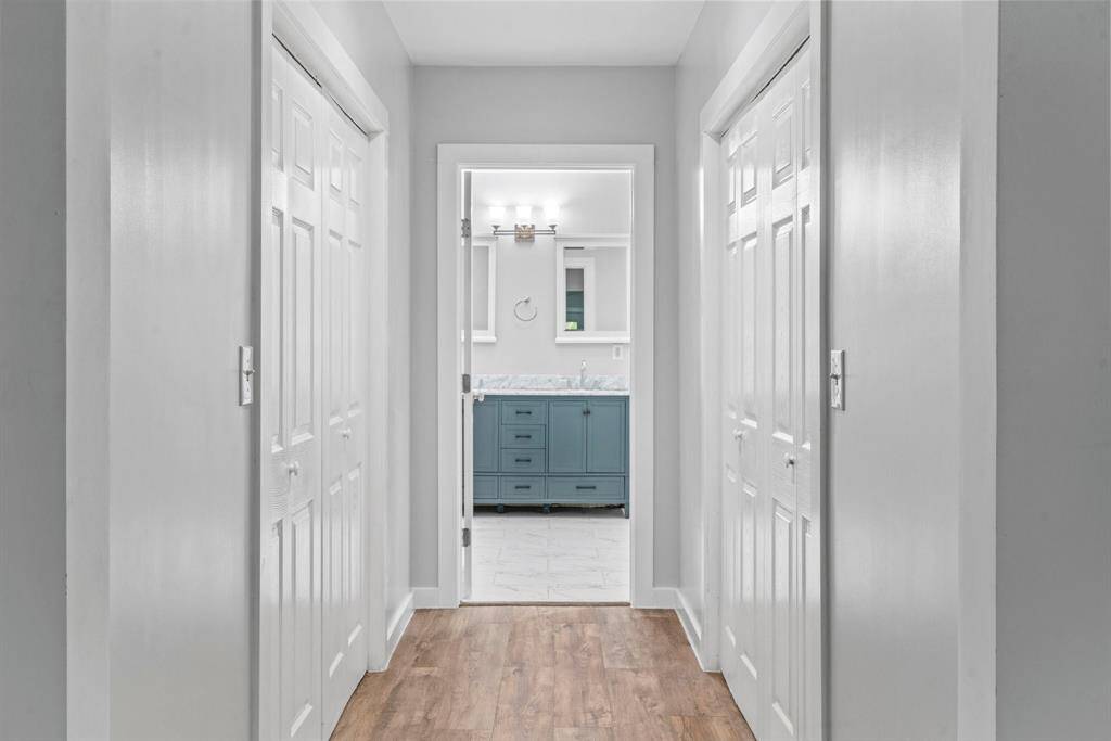 A bright hallway with wood flooring features dual white closet doors on both sides, leading to a modern bathroom with a blue vanity and large mirror.