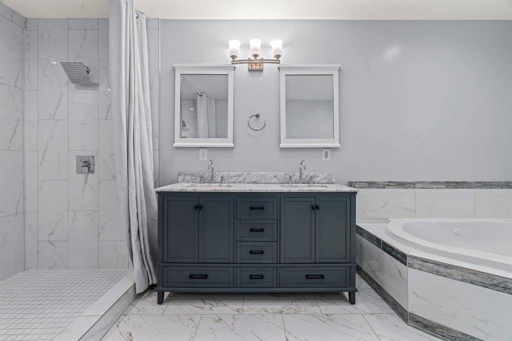 This modern bathroom features a sleek double vanity with marble countertops, a spacious walk-in shower with a rainfall showerhead, and a large soaking tub. The space is bright with tiled walls and floors, creating a luxurious and clean atmosphere.