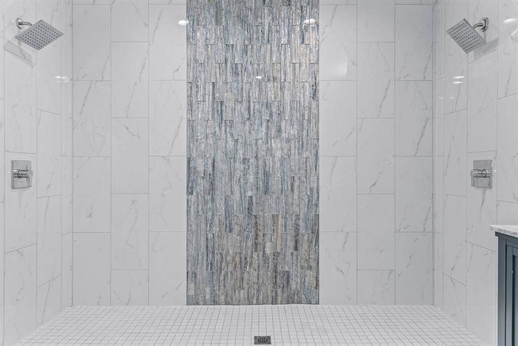 This bathroom features a spacious, modern shower with dual rainfall showerheads and elegant marble-like tiles, complemented by a striking vertical accent strip.