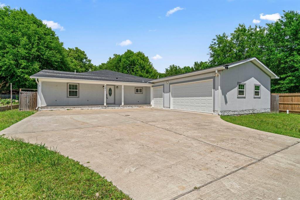 This photo shows a single-story gray house with a large driveway and a triple-car garage. The property has a well-maintained lawn and is surrounded by mature trees, offering a sense of privacy.