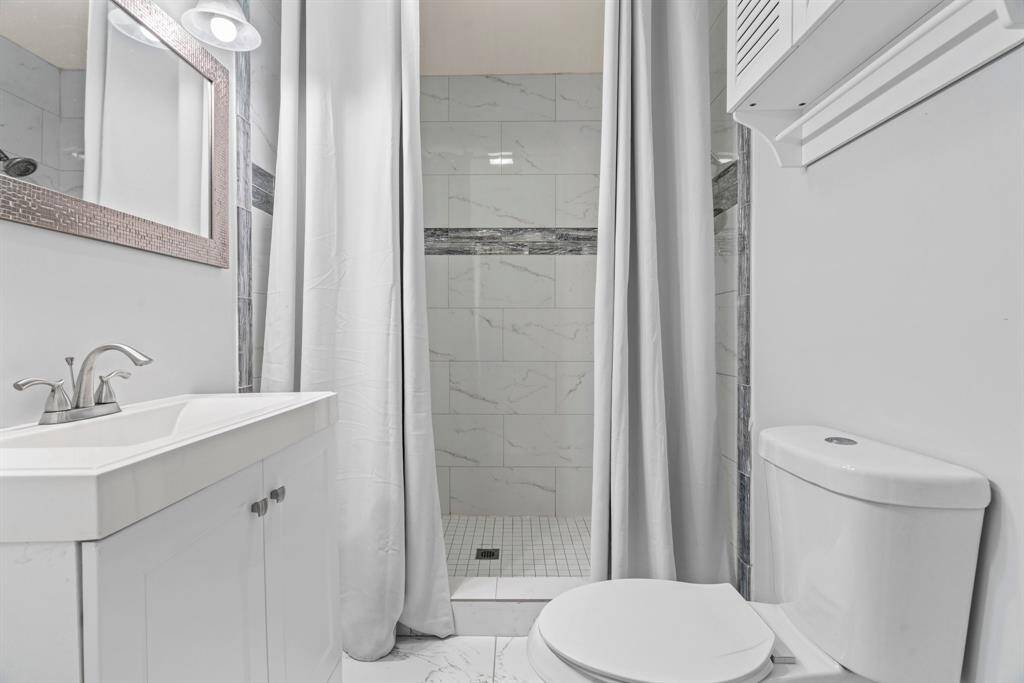 This bathroom features a modern, clean design with white tiles and fixtures, a sleek vanity with a mirrored cabinet, and a tiled walk-in shower with a curtain.