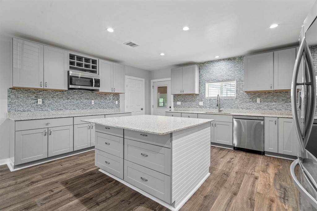 This kitchen features modern gray cabinetry, a spacious island, stainless steel appliances, and a stylish tile backsplash. The open layout and ample counter space make it ideal for cooking and entertaining. The wood flooring adds warmth and elegance.