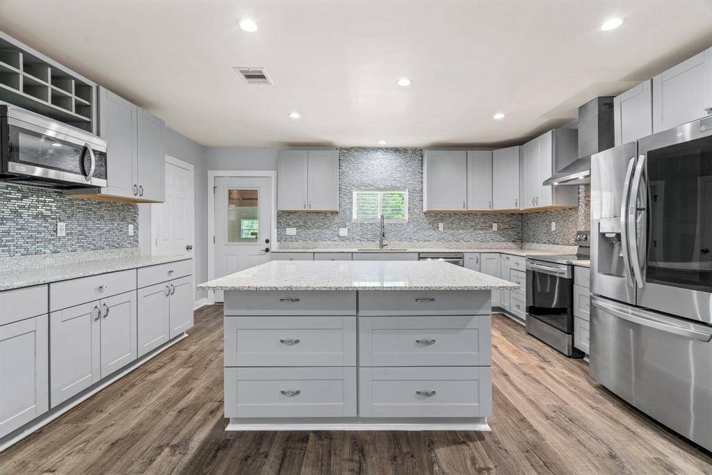 This kitchen features modern gray cabinetry, stainless steel appliances, and a large island with granite countertops. The backsplash is a sleek mosaic tile, and the room is well-lit with recessed lighting, creating a bright and inviting space. The wood flooring adds warmth and elegance.