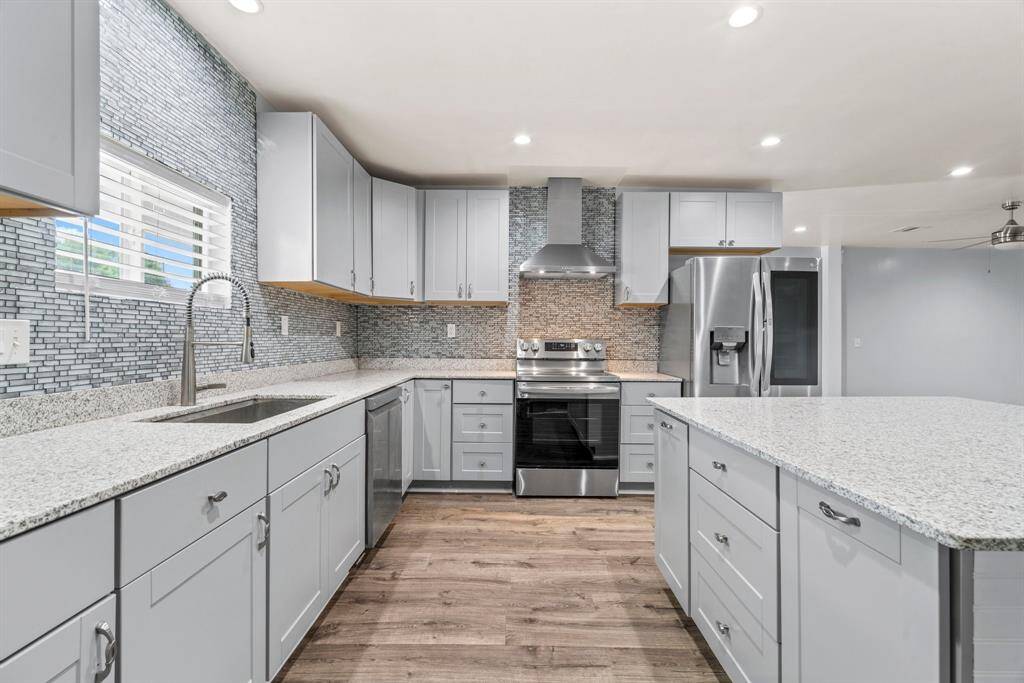 This modern kitchen features sleek gray cabinetry, granite countertops, and stainless steel appliances, including a double-door refrigerator and a built-in oven with a range hood. The space is well-lit with recessed lighting and has a stylish tile backsplash, complemented by the warm wood flooring.