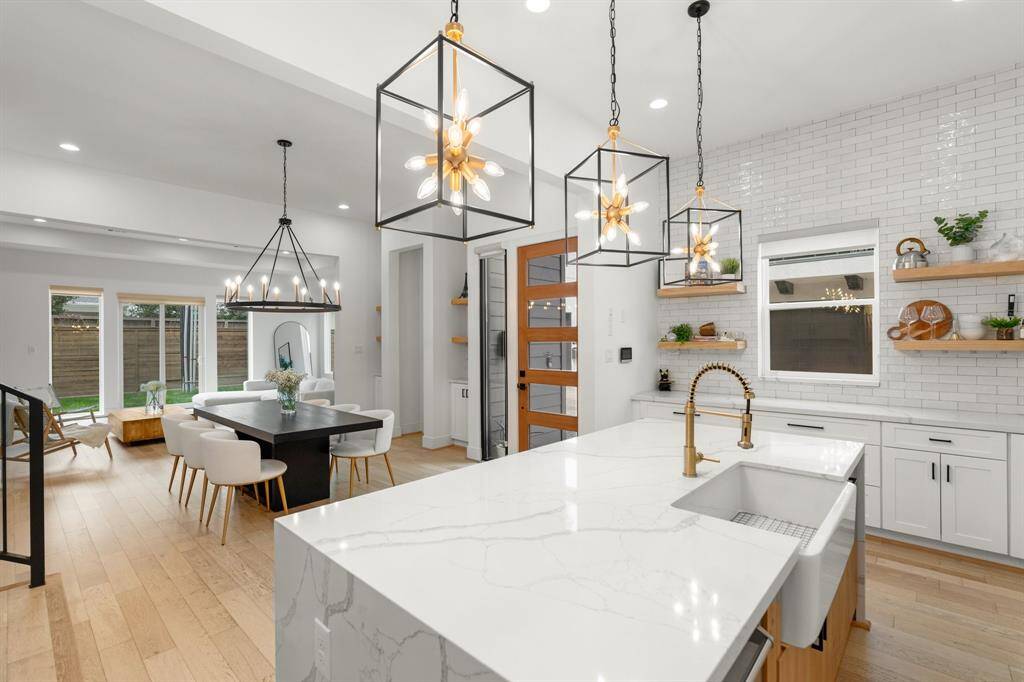 This home showcases a modern open-plan kitchen and dining area. It features a large island with a farmhouse sink, stylish pendant lighting, and white cabinetry. There's a spacious dining table with contemporary chairs, hardwood flooring, and ample natural light from large windows overlooking the backyard.