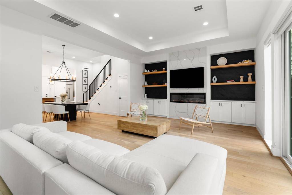 This modern living area features an open floor plan with a sleek fireplace, built-in shelving, and a spacious dining area. The room is well-lit with recessed lighting and natural light from large windows, offering a bright and inviting space.