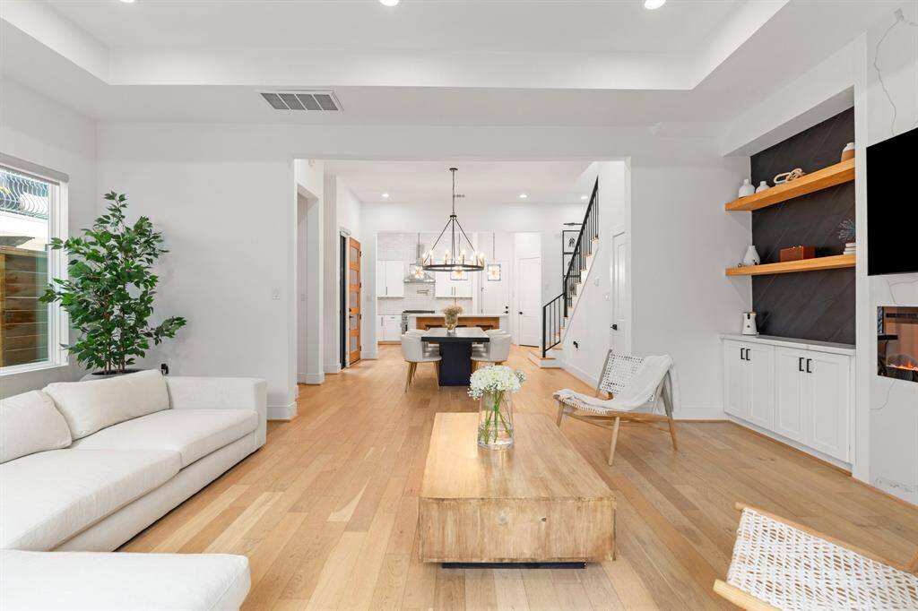 This photo features a modern, open-concept living and dining area with sleek wooden flooring. The space is bright and airy, featuring a cozy living area with stylish shelving. The dining area is highlighted by a contemporary chandelier, and the overall design is clean and inviting.